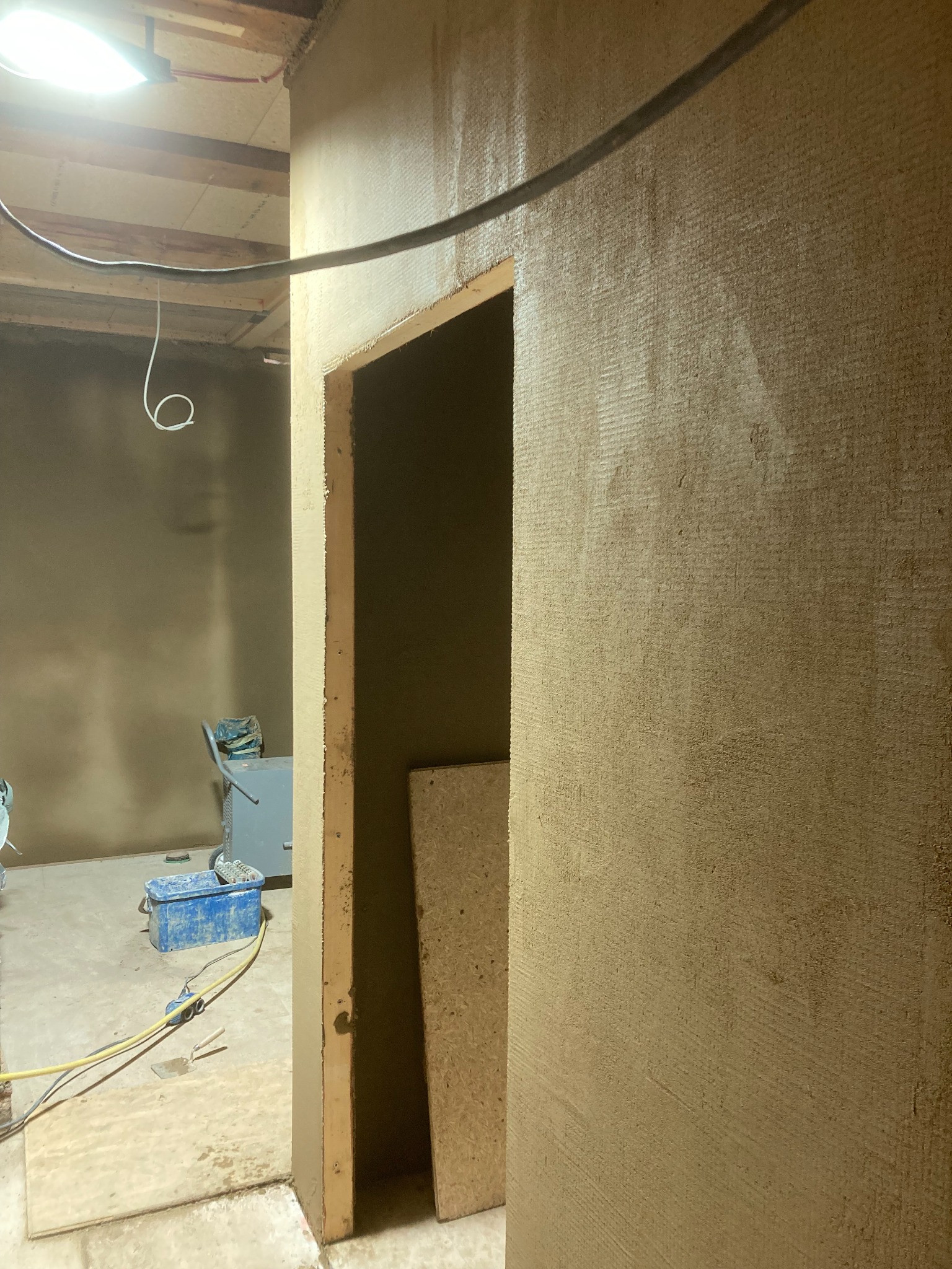 Świeżo nałożona warstwa mokrej zaprawy na ścianie w trakcie prac wykończeniowych, widoczne narzędzia na podłodze, niebieski pojemnik i maszyna do tynkowania w tle.