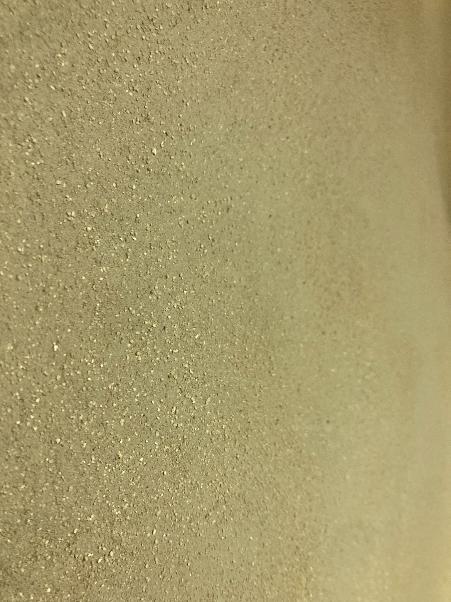 Szorstka powierzchnia ściany w odcieniach beżu i szarości, z widoczną strukturą drobnych ziarenek, oświetlona z prawej strony, tworząc delikatny cień.