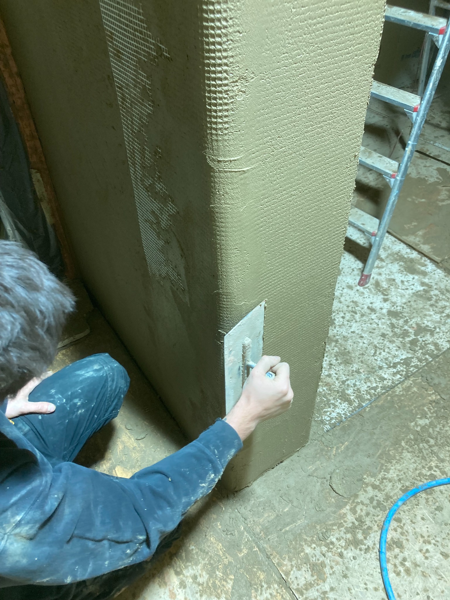 Pracownik nakłada zaprawę na ścianę wzmocnioną siatką, widoczna drabina i zabrudzone podłoże.