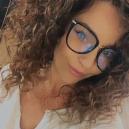 Portret kobiety z kręconymi włosami, noszącej okulary w czarnych oprawkach, ubranej w białą koszulę.