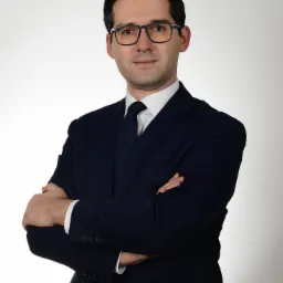 Portret mężczyzny w okularach, granatowym garniturze i krawacie, z założonymi rękami na tle jasnego studia.