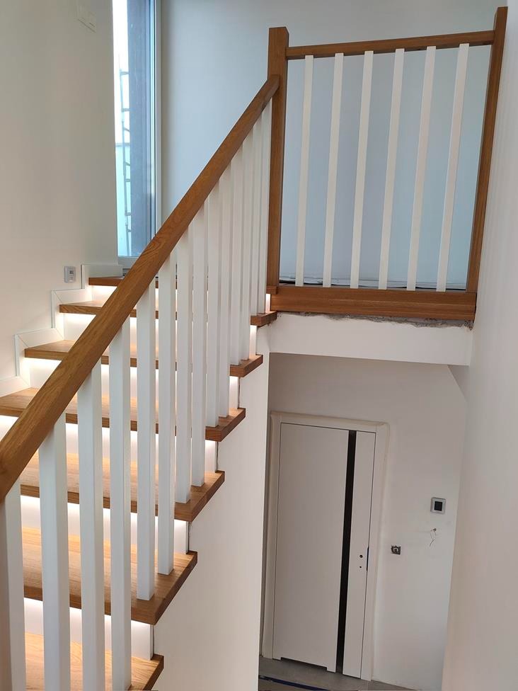 Nowoczesne schody z drewnianymi stopniami podświetlonymi od spodu, białe balustrady z drewnianym poręczem i dodatkową barierką zabezpieczającą na podeście, widok z góry na dół.