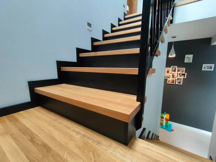 Nowoczesne schody z drewnianymi stopniami i czarną konstrukcją, widok z dołu ukazujący fragment podłogi i balustradę z czarnymi prętami.