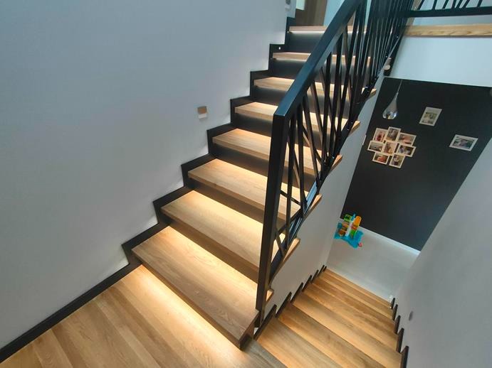 Nowoczesne schody drewniane z podświetleniem LED i czarną, geometryczną balustradą, widok z góry na dół.