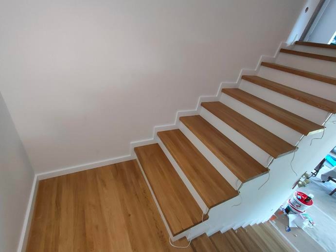 Nowoczesne, dębowe stopnie schodowe z białymi podstopnicami i listwami przypodłogowymi, widoczne ślady po malowaniu na podłodze.