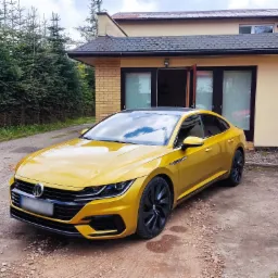 Żółty Volkswagen Arteon po modyfikacjach stoi przed domem w Słupsku, z czarnymi felgami i przyciemnionymi szybami.