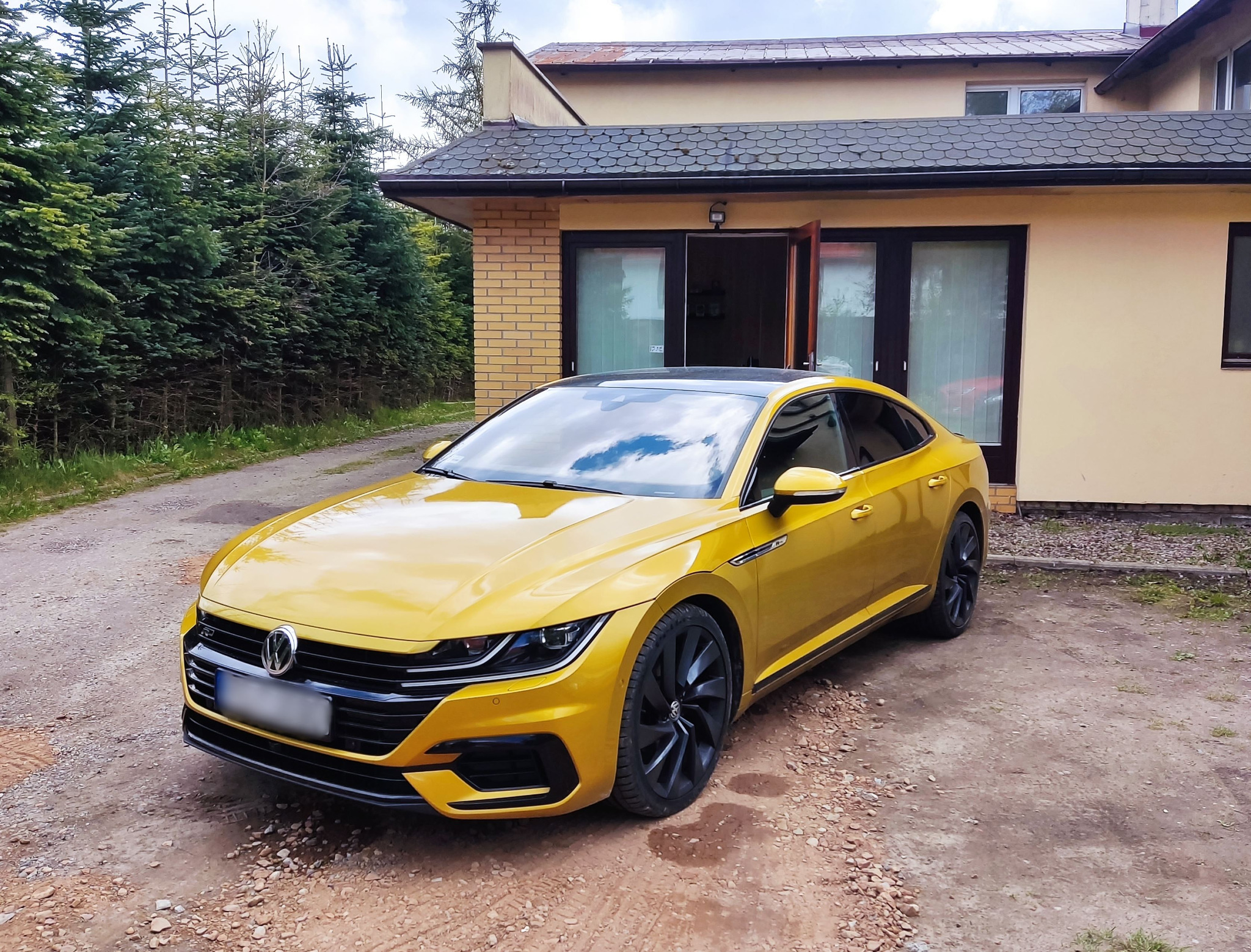 Żółty Volkswagen Arteon po modyfikacjach stoi przed domem w Słupsku, z czarnymi felgami i przyciemnionymi szybami.