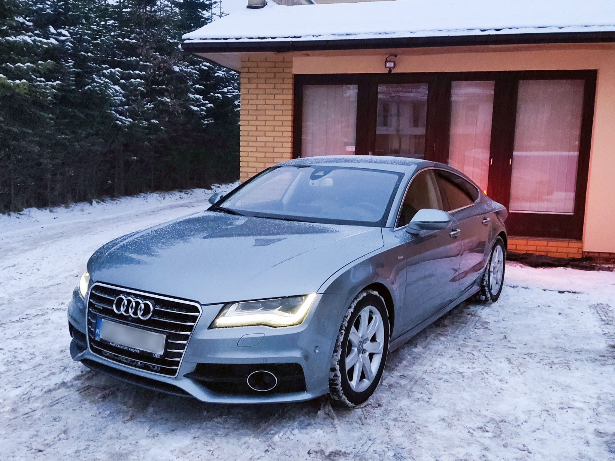 Szary samochód Audi A7 zaparkowany na śniegu przed domem w zimowej scenerii, z włączonymi światłami mijania i widocznymi detalami karoserii.