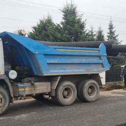F.H.U Gac - Ciężarówka Kamaz z niebieską skrzynią załadowana czarnymi rurami karbowanymi, obok koparka CAT i dwóch robotników w odblaskowych kamizelkach na tle budynków i drzew podczas robót drogowych.