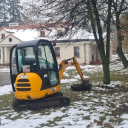 F.H.U Gac - Żółta mini koparka JCB 8018 w trakcie prac ziemnych na zaśnieżonym terenie, widoczny wykop przy drzewie i budynek w tle.
