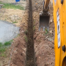 F.H.U Gac - Wąski wykop liniowy pod fundamenty domu jednorodzinnego, wykonany koparką JCB, z widocznym pracownikiem w tle i niebieskim wężem doprowadzającym wodę.