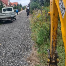 F.H.U Gac - Żwirowa droga w trakcie utwardzania, widok z kabiny koparki, pracownicy porządkują teren, w tle zabudowa jednorodzinna i dostawczy samochód.