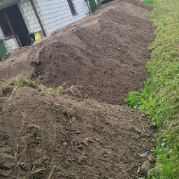 F.H.U Gac - Nierówna pryzma ciemnej ziemi usypana wzdłuż budynku z białej cegły i zielonego trawnika, w tle niebieska wywrotka.