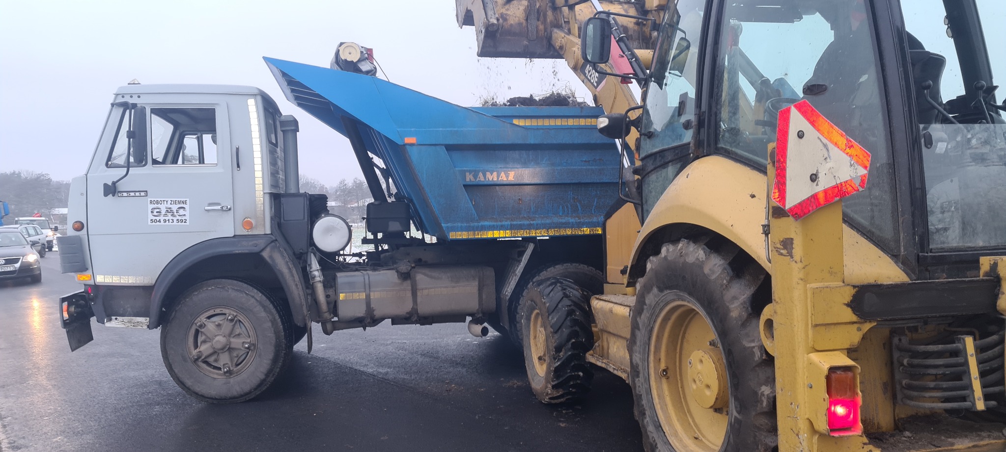 Koparko-ładowarka z łyżką wysypuje ziemię do niebieskiej wywrotki Kamaz na tle mglistego krajobrazu.