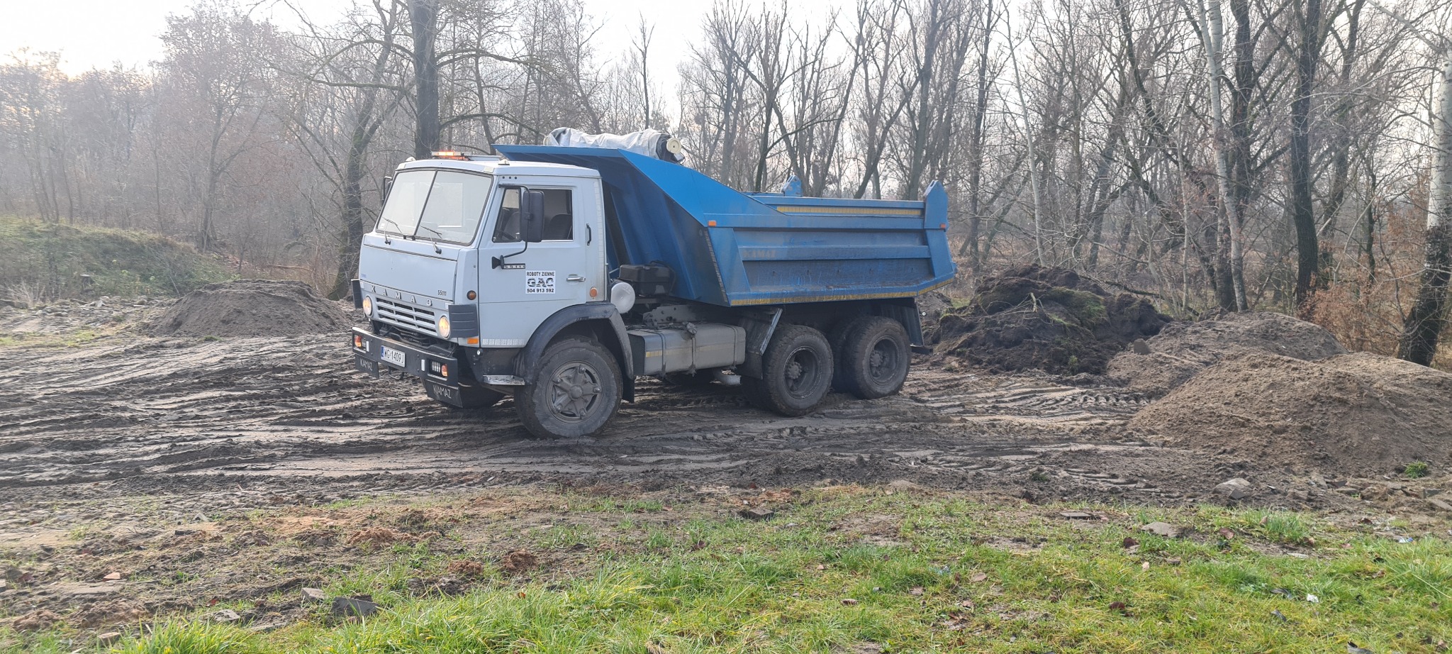 Ciężarówka wywrotka Kamaz na błotnistym terenie z pryzmami ziemi w tle, widoczne ślady opon i tablica rejestracyjna WG 1409J.
