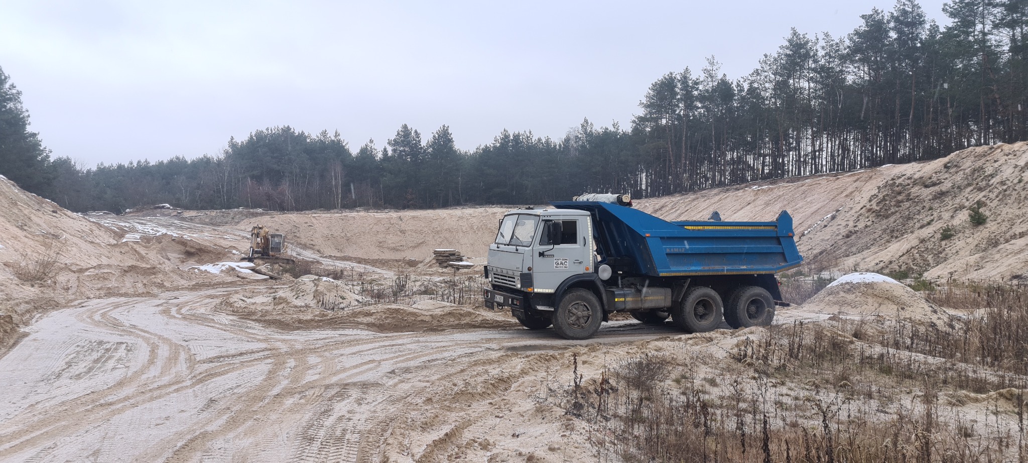 Ciężarówka wywrotka KAMAZ z niebieską skrzynią ładunkową na piaszczystym placu budowy z widoczną koparką w tle, otoczona lasem iglastym przy pochmurnej pogodzie.