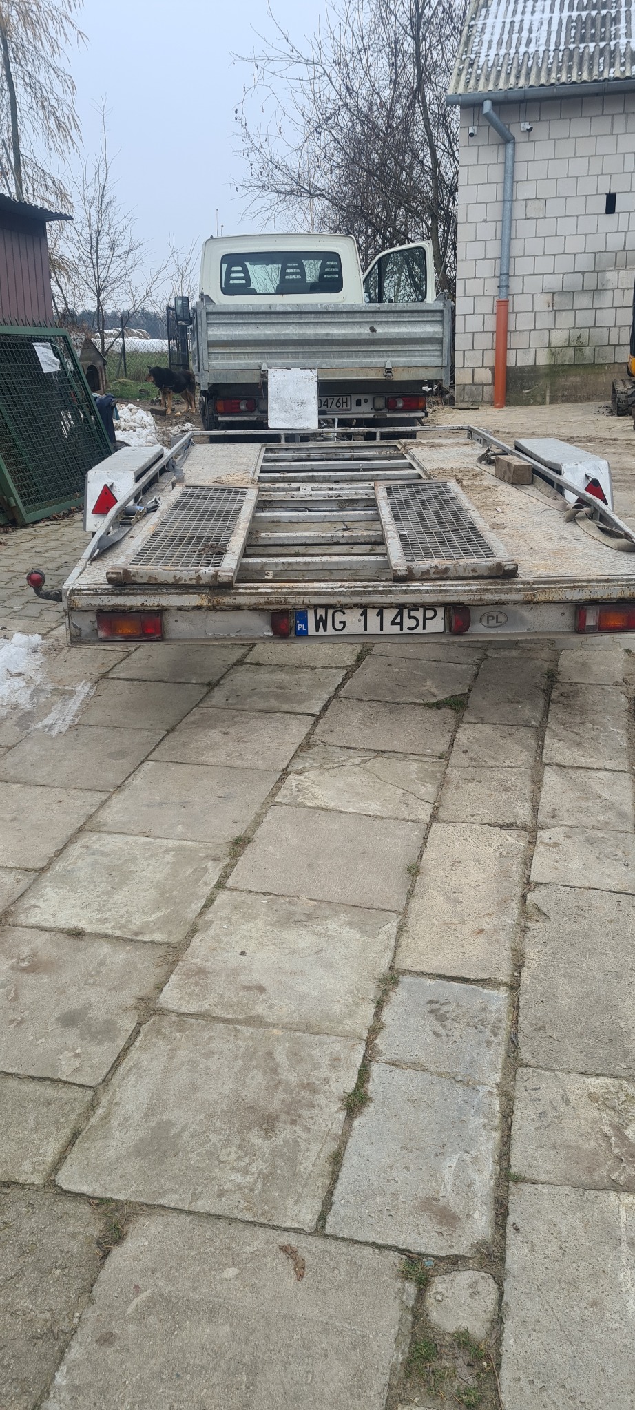 Przyczepa samochodowa z metalową kratownicą na betonowej nawierzchni z widocznym samochodem dostawczym w tle.