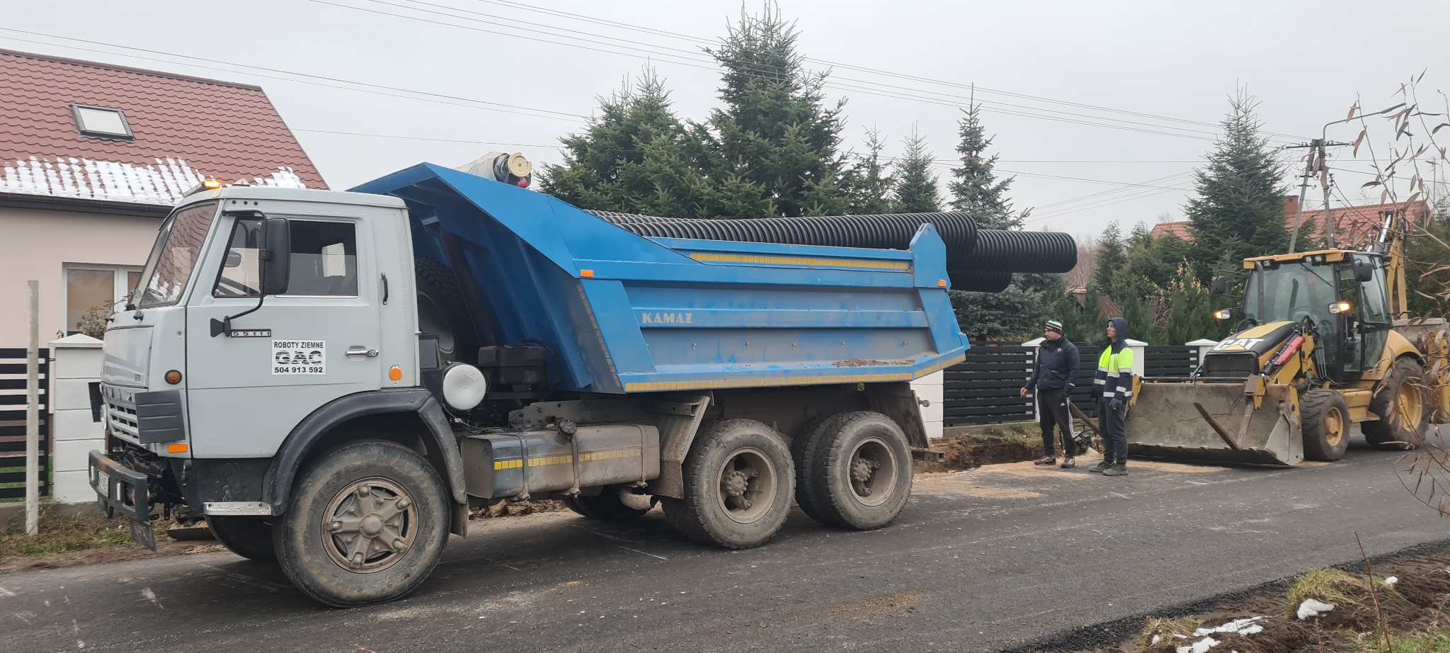 Ciężarówka Kamaz z niebieską skrzynią załadowana czarnymi rurami karbowanymi, obok koparka CAT i dwóch robotników w odblaskowych kamizelkach na tle budynków i drzew podczas robót drogowych.