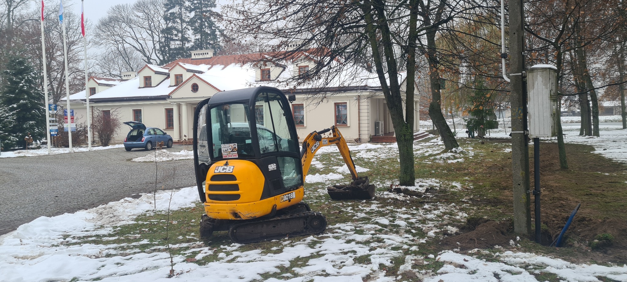 Żółta mini koparka JCB 8018 w trakcie prac ziemnych na zaśnieżonym terenie, widoczny wykop przy drzewie i budynek w tle.