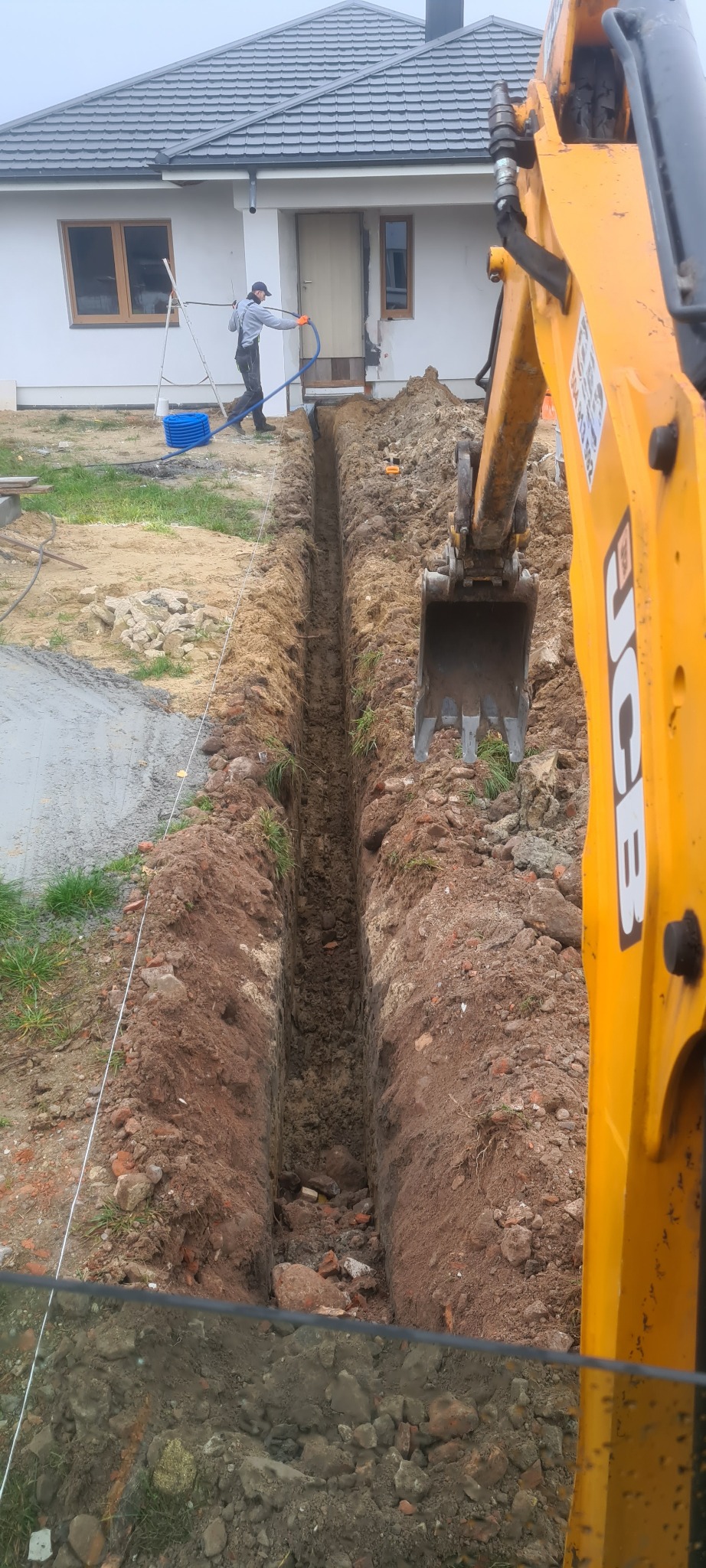 Wąski wykop liniowy pod fundamenty domu jednorodzinnego, wykonany koparką JCB, z widocznym pracownikiem w tle i niebieskim wężem doprowadzającym wodę.