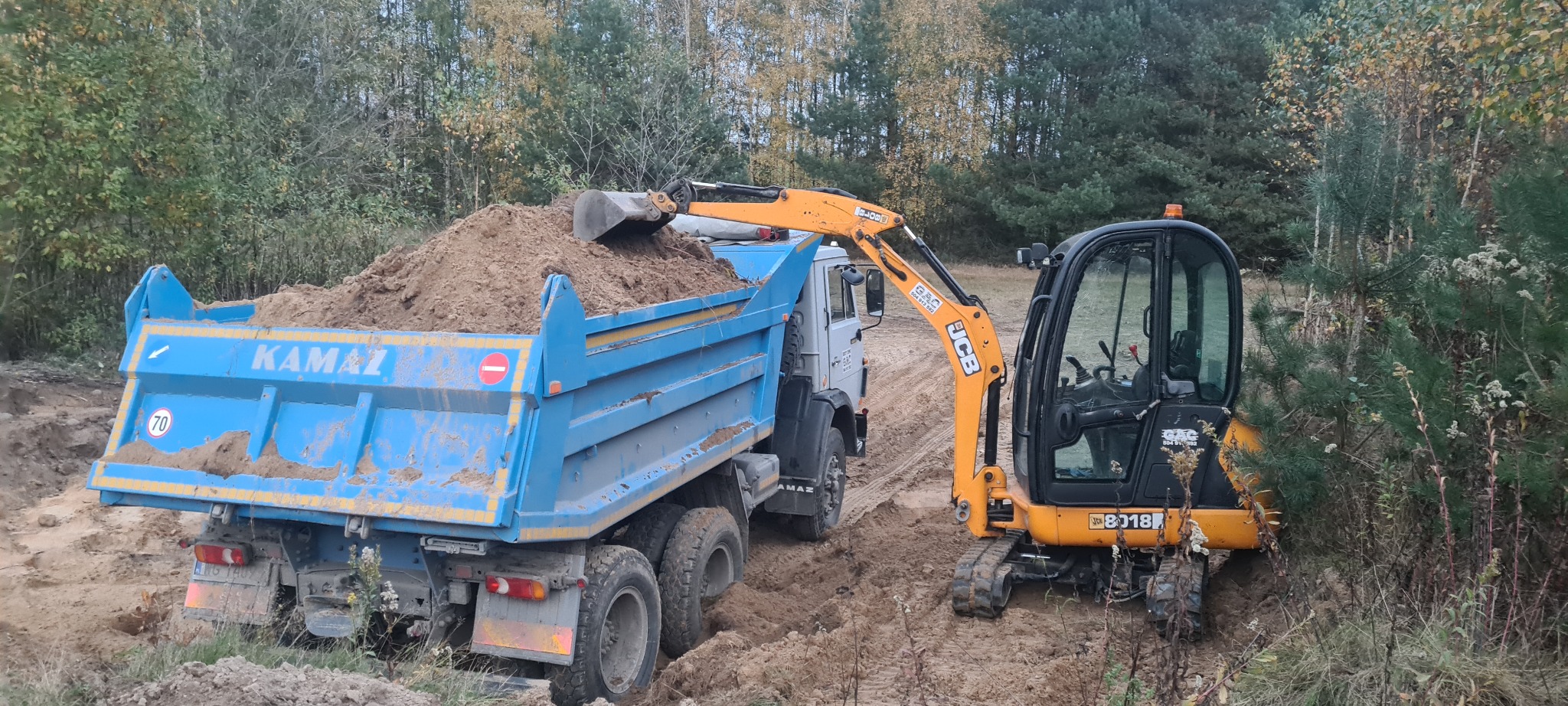 Żółta koparka JCB ładuje ziemię do niebieskiej ciężarówki Kamaz na terenie budowy otoczonym drzewami.