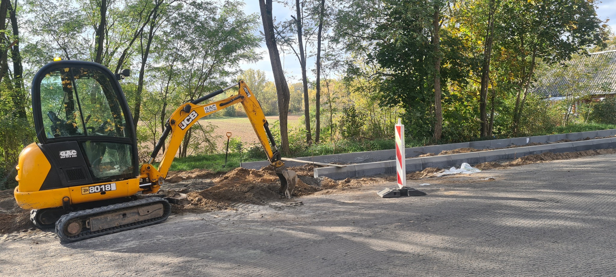 Żółta mini koparka JCB 8018 przygotowuje teren pod krawężniki na nowej drodze asfaltowej, w tle drzewa i budynek.
