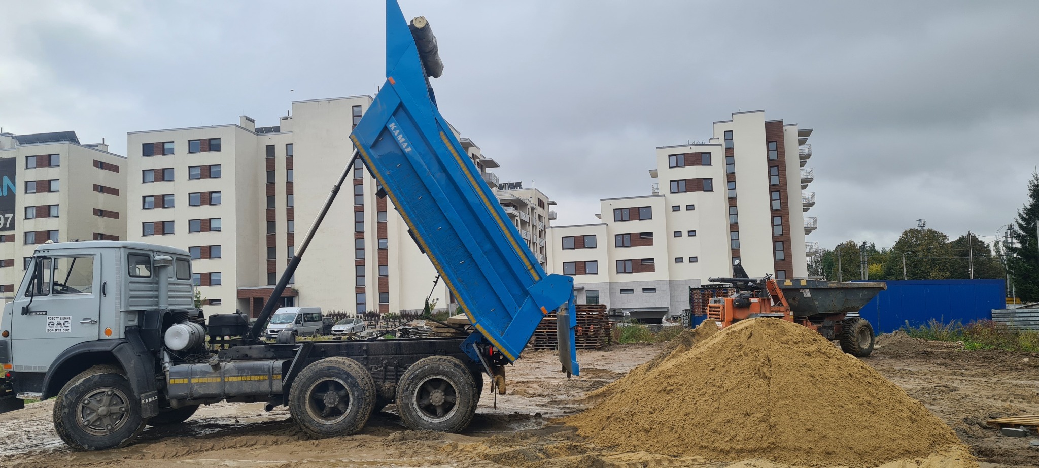 Ciężarówka Kamaz z podniesioną, niebieską skrzynią ładunkową wysypuje piasek na placu budowy, w tle budynki mieszkalne.