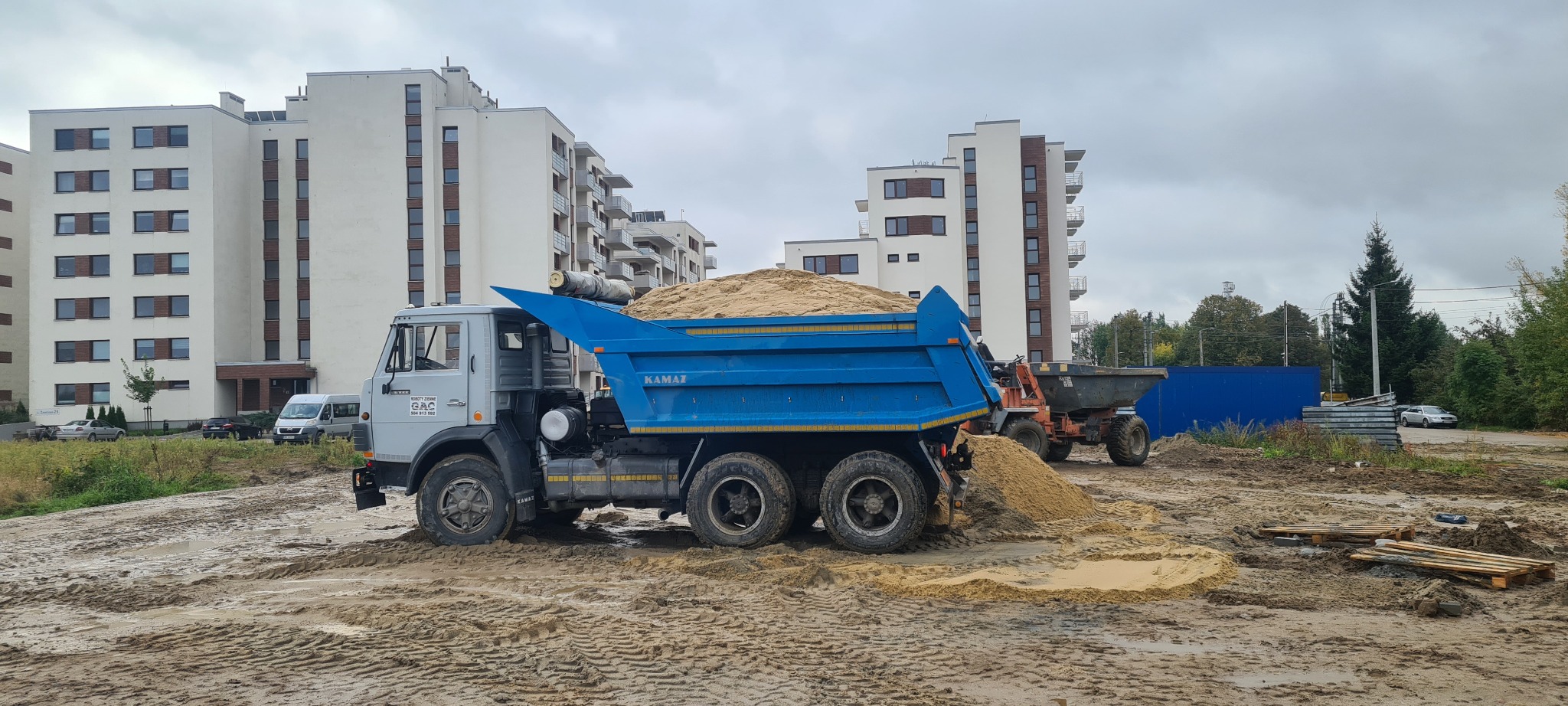 Ciężarówka KAMAZ z niebieską skrzynią załadowana piaskiem na placu budowy, w tle białe budynki mieszkalne i błotniste podłoże.