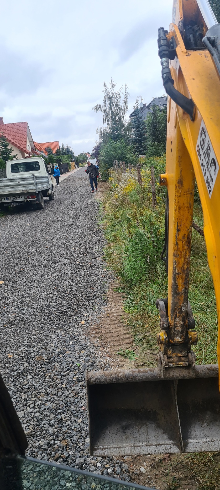 Żwirowa droga w trakcie utwardzania, widok z kabiny koparki, pracownicy porządkują teren, w tle zabudowa jednorodzinna i dostawczy samochód.