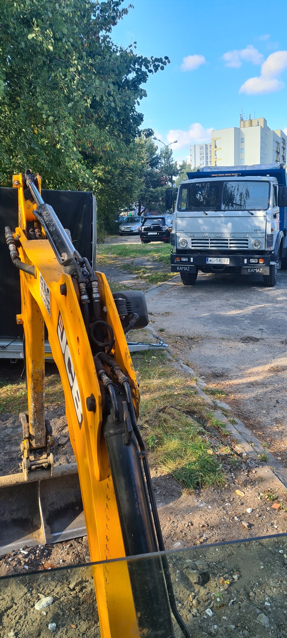 Żółta koparko-ładowarka JCB na placu budowy, z wykopem w tle, obok stoi wywrotka Kamaz i zaparkowane samochody, w tle budynki mieszkalne i drzewa.
