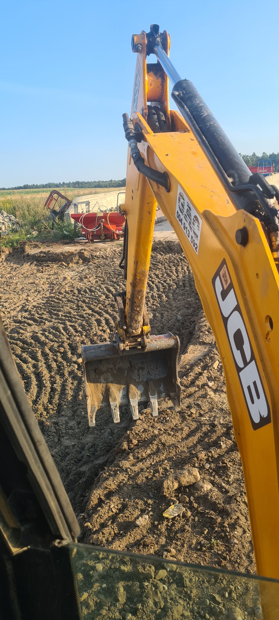 Żółta koparka JCB z łyżką zagłębiającą się w ziemię, widok z kabiny, w tle rozrzucone kamienie i maszyny rolnicze pod błękitnym niebem.