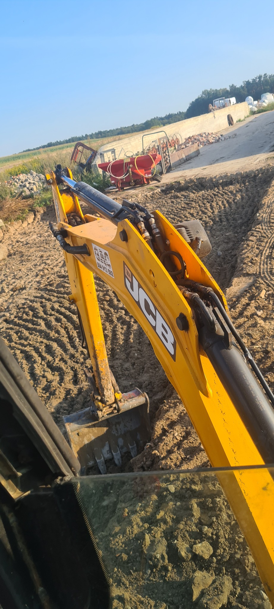 Żółta koparka JCB w trakcie prac ziemnych, widok z kabiny operatora, w tle sprzęt rolniczy i składowane materiały budowlane.