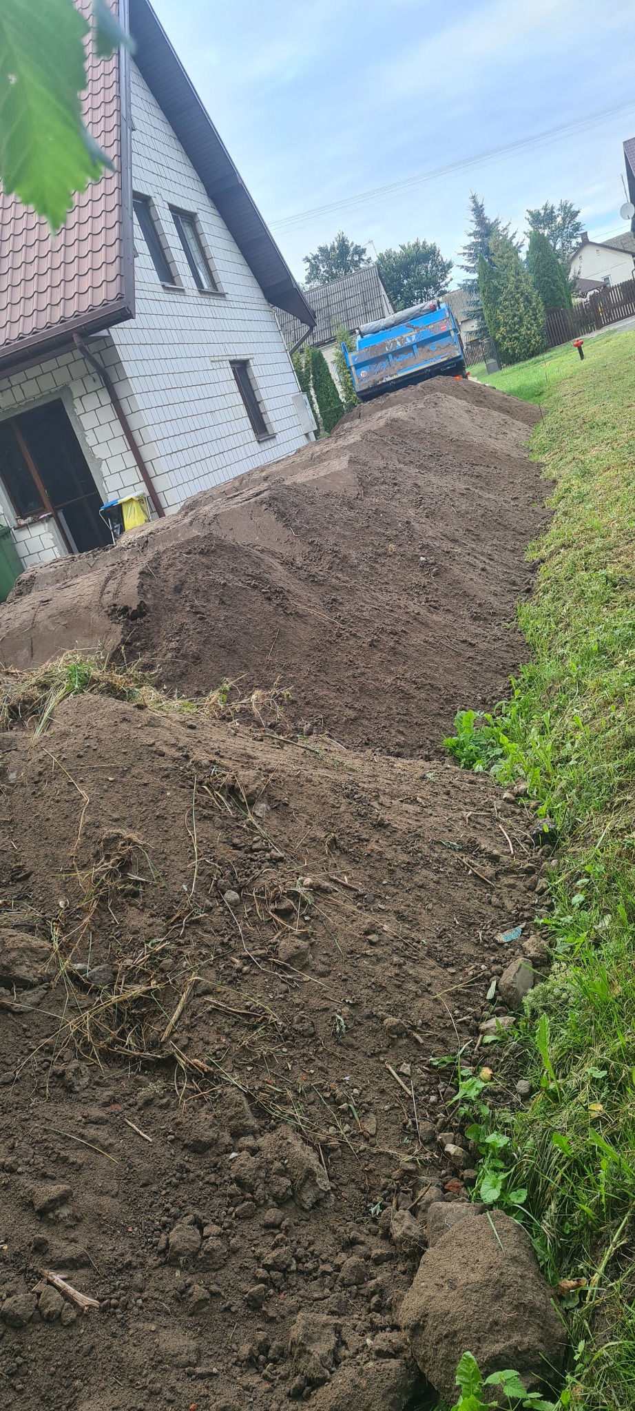 Nierówna pryzma ciemnej ziemi usypana wzdłuż budynku z białej cegły i zielonego trawnika, w tle niebieska wywrotka.