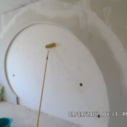 MĘŻCZYZNA w domu - Półokrągła wnęka w ścianie w trakcie szpachlowania, widoczny wałek malarski oparty o ścianę, worek z materiałem budowlanym w rogu pomieszczenia, surowy stan wykończenia.