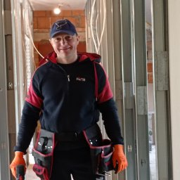 MĘŻCZYZNA w domu - Uśmiechnięty monter w roboczym stroju i pomarańczowych rękawicach, stojący wewnątrz konstrukcji ścian działowych z profili metalowych, w tle widoczny surowy mur z cegły.