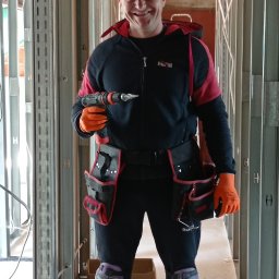 MĘŻCZYZNA w domu - Uśmiechnięty pracownik w stroju roboczym, z wiertarką w dłoni, stoi wewnątrz konstrukcji ścian działowych z metalowych profili, widoczne instalacje elektryczne.