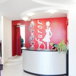 MĘŻCZYZNA w domu - Recepcja salonu z czerwonym akcentem na ścianie, białym napisem 'Furor' i sylwetką kobiety, za ladą recepcji osoba obsługująca klientów, po lewej manekin w fioletowej sukience.