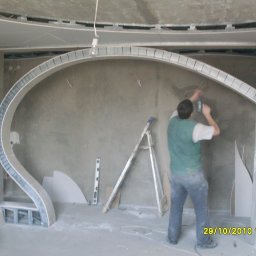 MĘŻCZYZNA w domu - Pracownik montuje metalową konstrukcję łukową z płyt kartonowo-gipsowych na ścianie, widoczna drabina i poziomica, surowy stan wykończenia wnętrza.