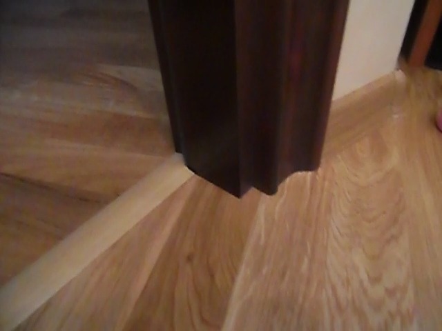 Fragment podłogi z jasnego drewna, widoczna listwa przypodłogowa i ciemna framuga drzwi, ujęcie z niskiego kąta.