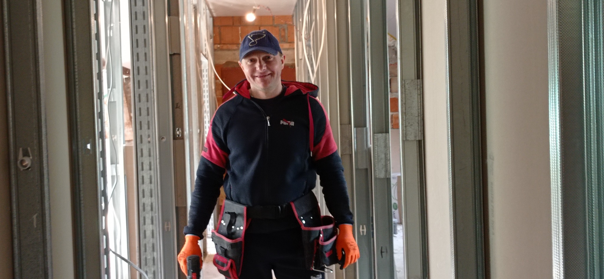 Uśmiechnięty monter w roboczym stroju i pomarańczowych rękawicach, stojący wewnątrz konstrukcji ścian działowych z profili metalowych, w tle widoczny surowy mur z cegły.