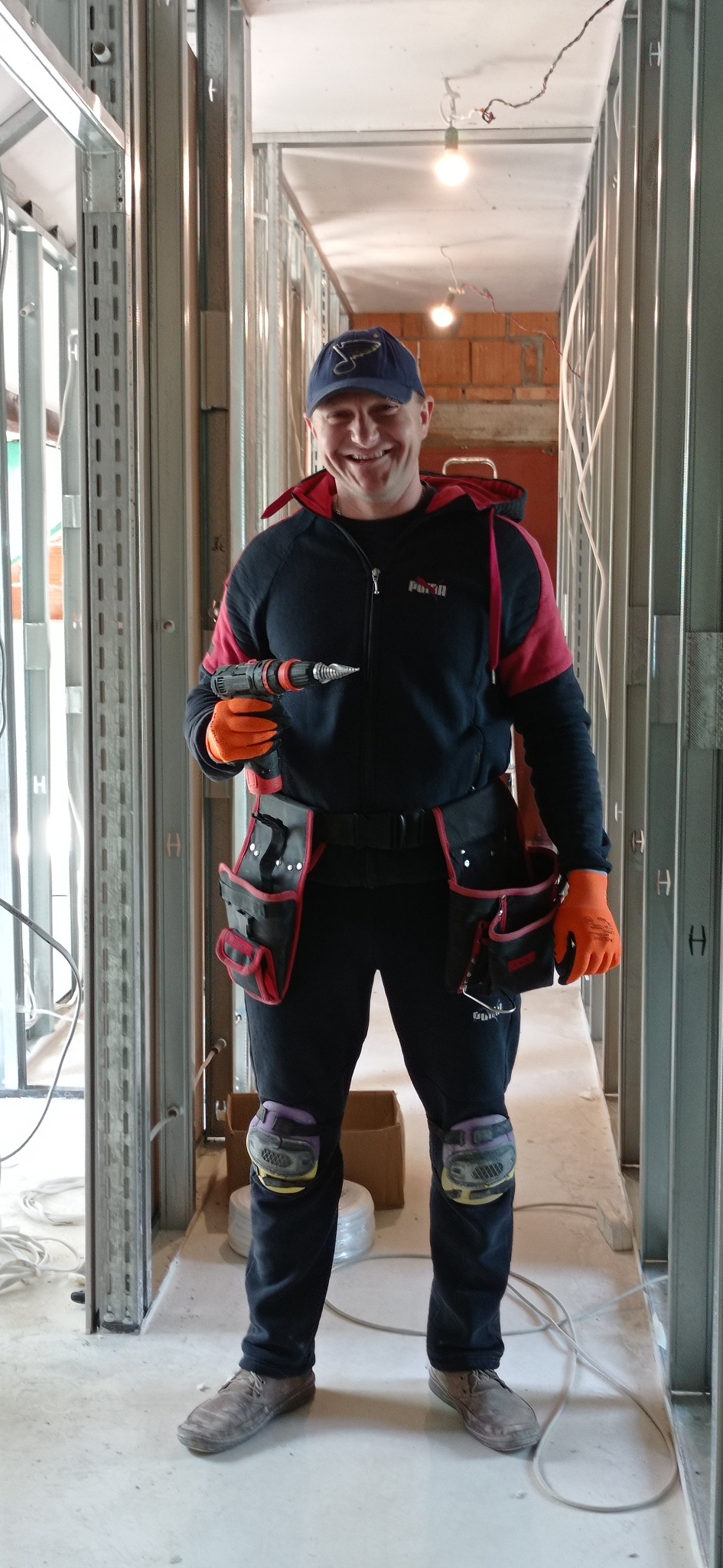 Uśmiechnięty pracownik w stroju roboczym, z wiertarką w dłoni, stoi wewnątrz konstrukcji ścian działowych z metalowych profili, widoczne instalacje elektryczne.