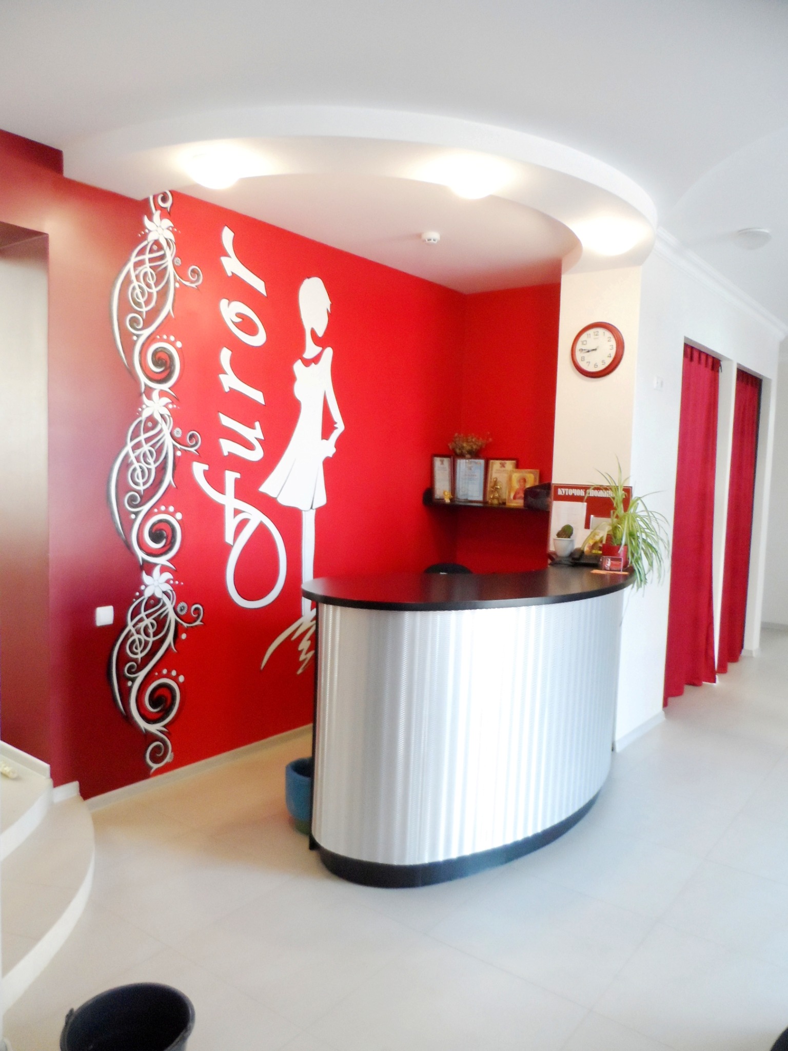 Recepcja z czerwonymi ścianami, dekoracyjnym ornamentem i sylwetką kobiety, okrągły blat, zegar na ścianie i czerwone zasłony w tle.