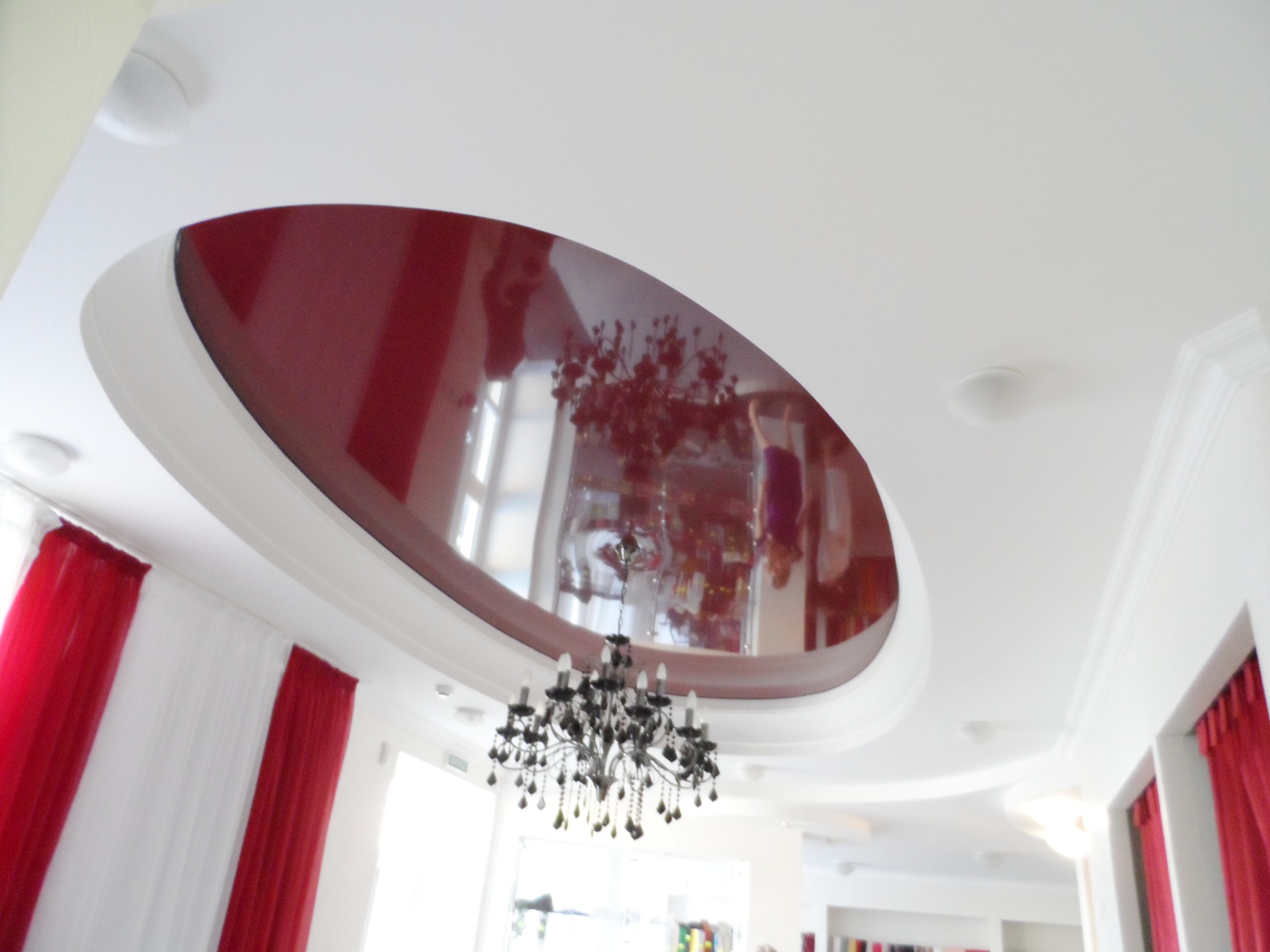 Błyszczący, bordowy sufit podwieszany o owalnym kształcie, odbijający światło i elementy wystroju, z widocznym żyrandolem z kryształkami, na tle białych ścian i czerwono-białych zasłon.