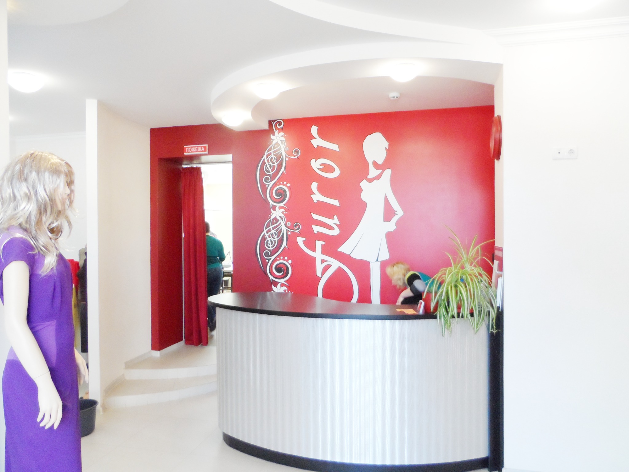 Recepcja salonu z czerwonym akcentem na ścianie, białym napisem 'Furor' i sylwetką kobiety, za ladą recepcji osoba obsługująca klientów, po lewej manekin w fioletowej sukience.