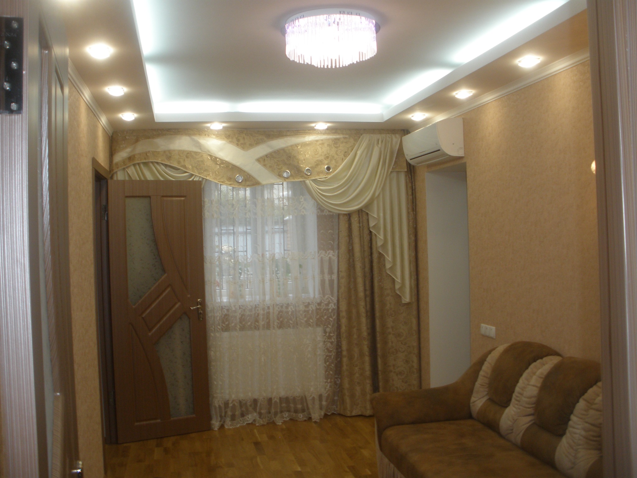 Wykończone wnętrze pokoju z dekoracyjnymi zasłonami, kremowymi tapetami, podwieszanym sufitem z oświetleniem punktowym i ozdobnym żyrandolem, widoczny fragment brązowej sofy.