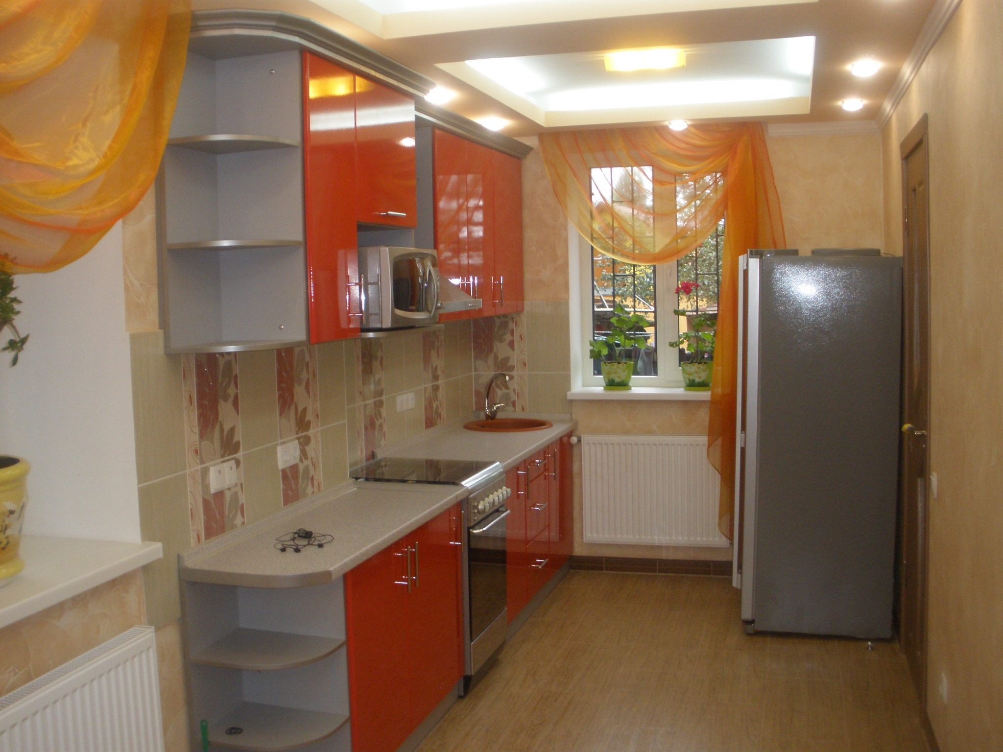 Jasna kuchnia z pomarańczowymi akcentami: błyszczące fronty szafek, beżowe płytki z motywem roślinnym, okno z firanką i widokiem na zieleń.