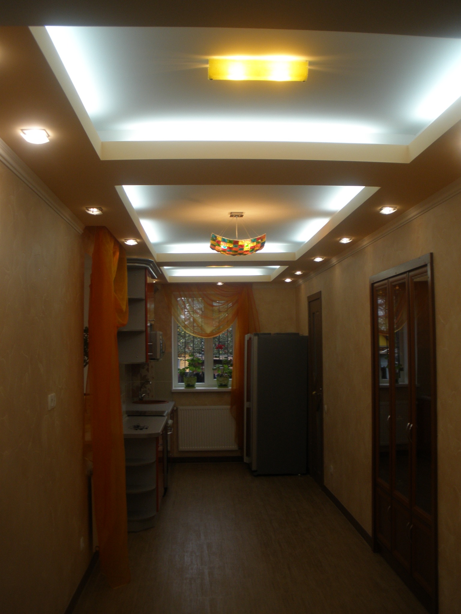 Wnętrze wąskiego korytarza z widokiem na kuchnię, beżowe ściany z delikatną fakturą, podwieszany sufit z oświetleniem LED i punktowymi lampami, parkiet imitujący drewno, okno z pomarańczową firanką.