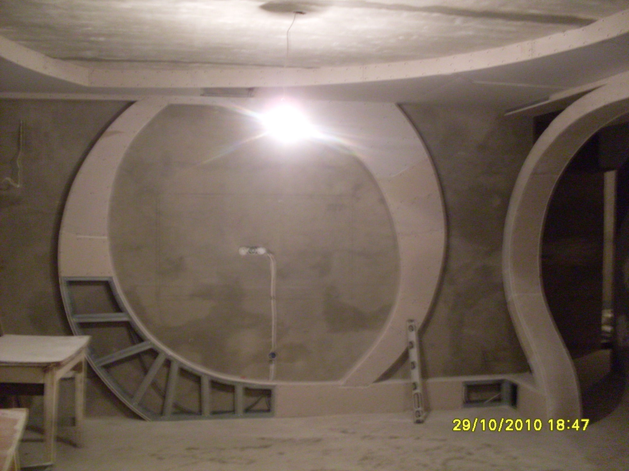 Niedokończona konstrukcja z płyt gipsowo-kartonowych w kształcie okręgu na ścianie, widoczny stelaż, poziomica i lampa halogenowa, surowy stan wykończenia wnętrza.