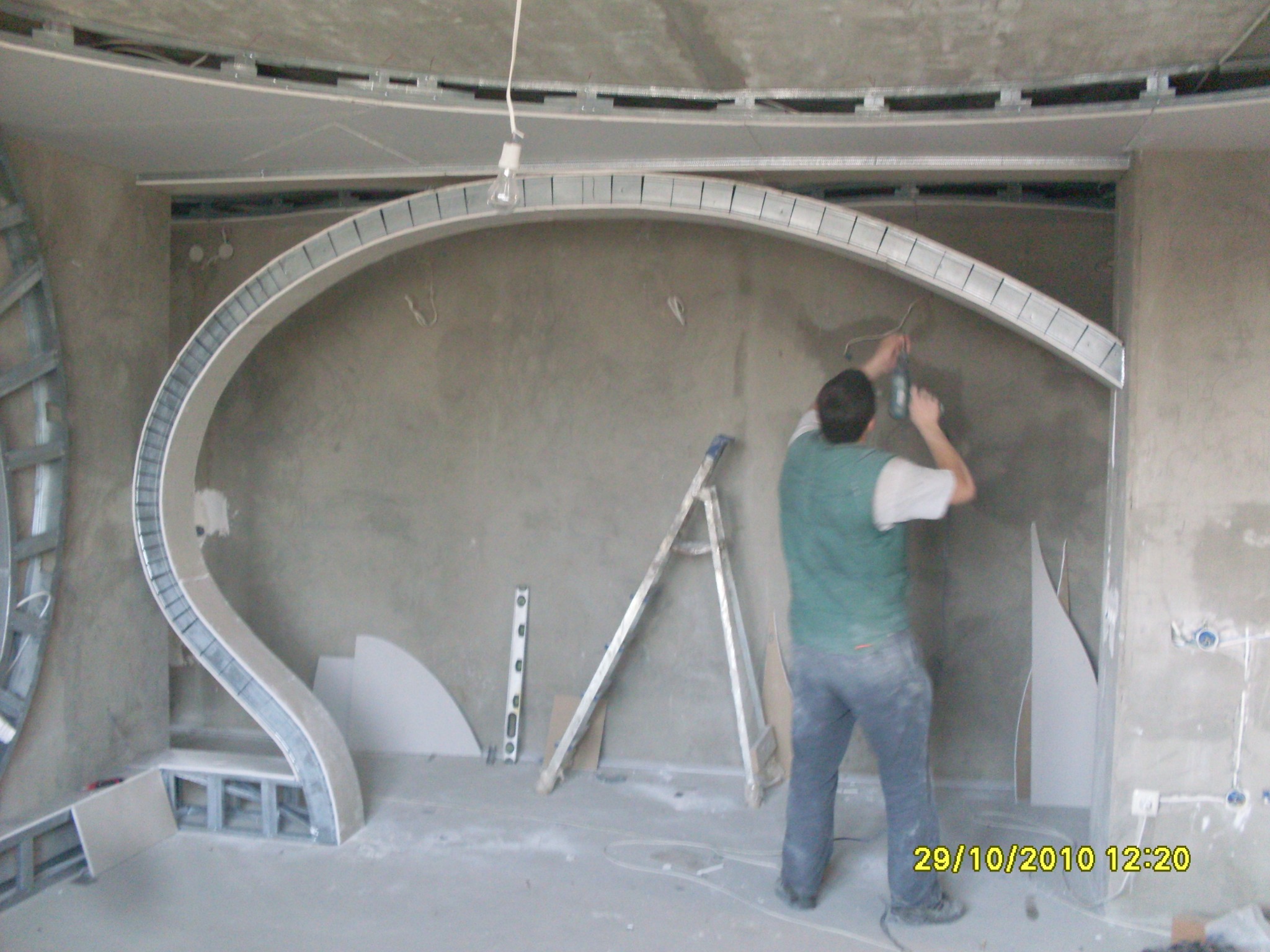 Pracownik montuje metalową konstrukcję łukową z płyt kartonowo-gipsowych na ścianie, widoczna drabina i poziomica, surowy stan wykończenia wnętrza.