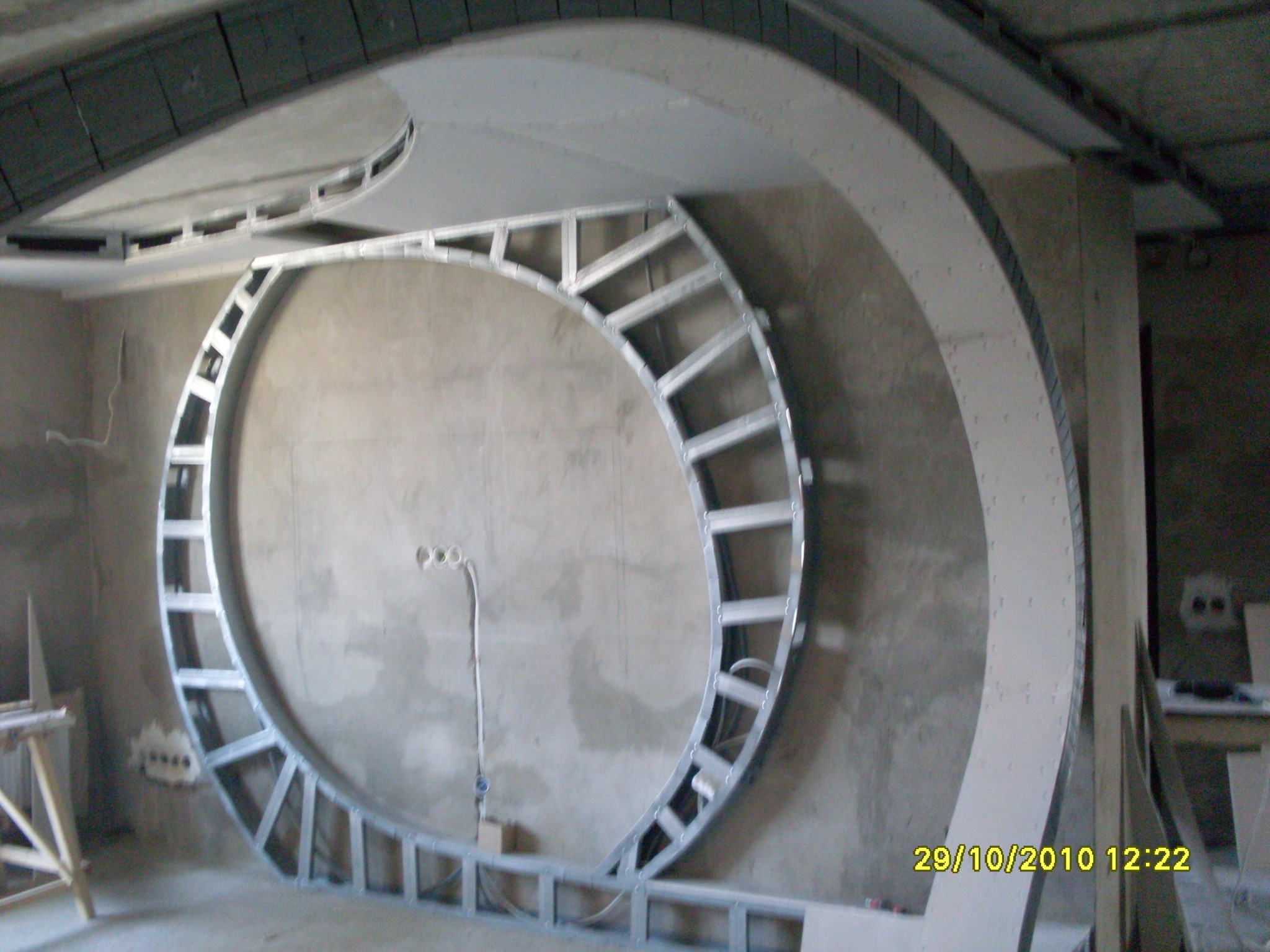 Niedokończona konstrukcja sufitu podwieszanego z metalowych profili w kształcie okręgu i łuku, widoczne przewody elektryczne, surowy stan wnętrza.